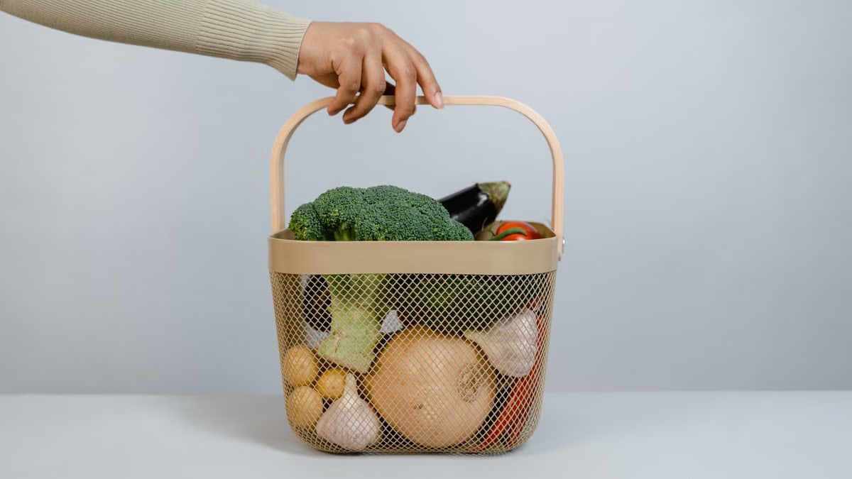 A hand holding a basket filled with fresh vegetables like broccoli and onions.