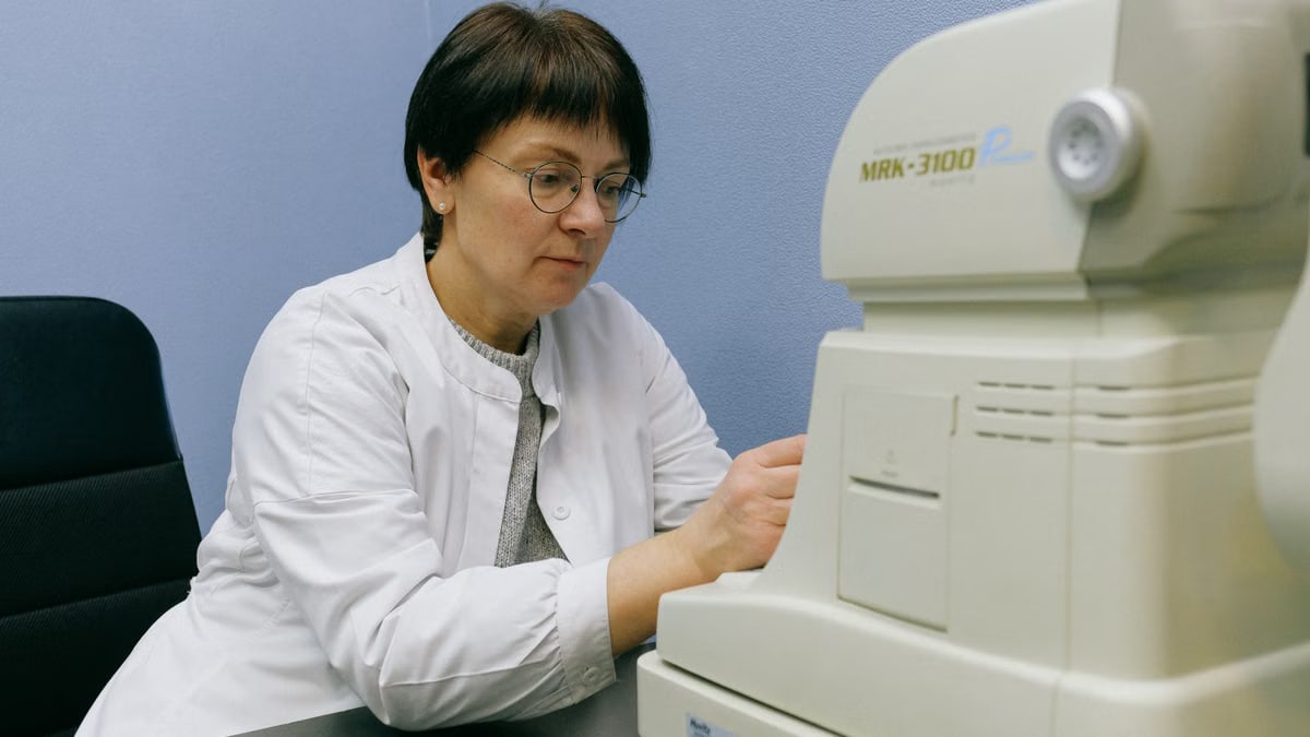 Serious middle aged eye doctor in eyeglasses working with automatic refkeratometer while sitting on chair leaned forward at black desk at work